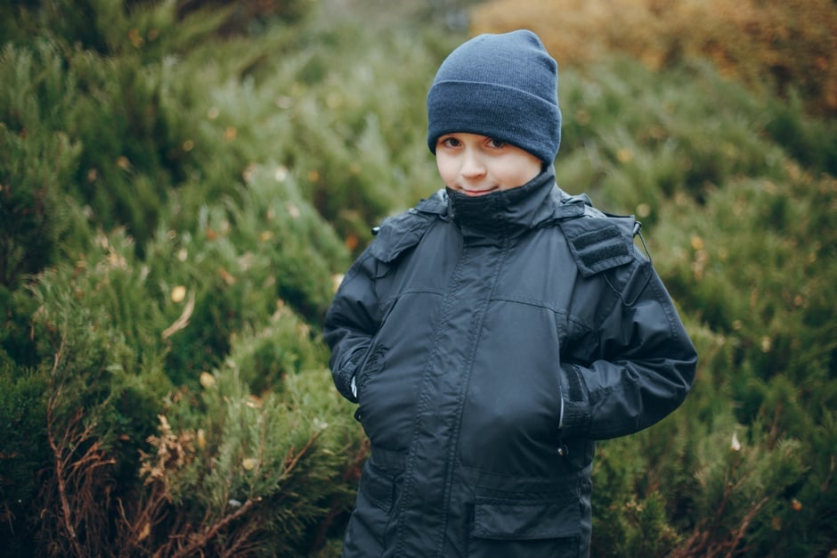 A clear picture of a boy wearing a black beanie and black jacket. Standing in front of a set of bushes.